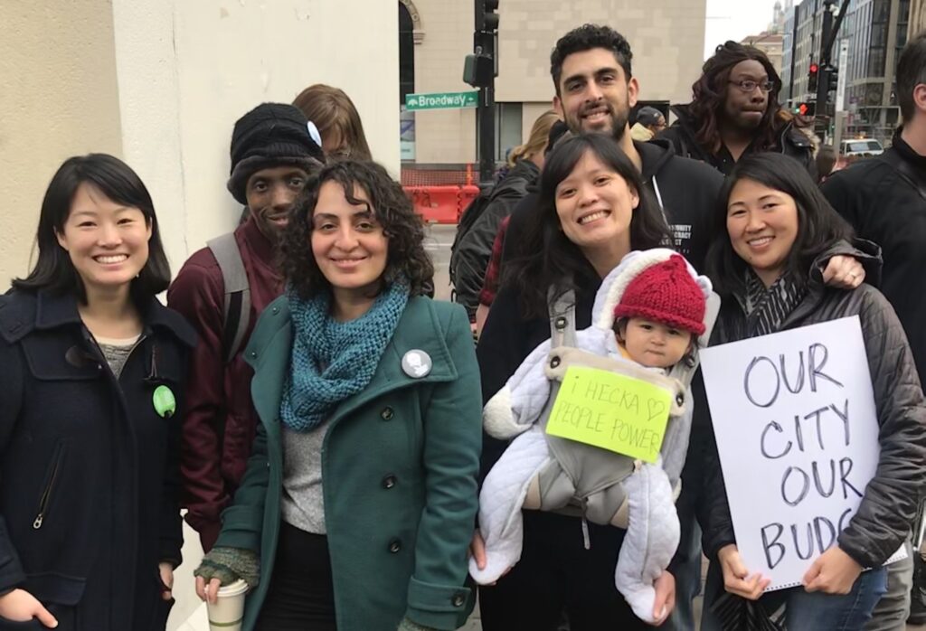 CDP members and supports smiling together at a protest in downtown Oakland. Baby wearing a baby bjorn holds a sign saying “I Hecka Love People Power”